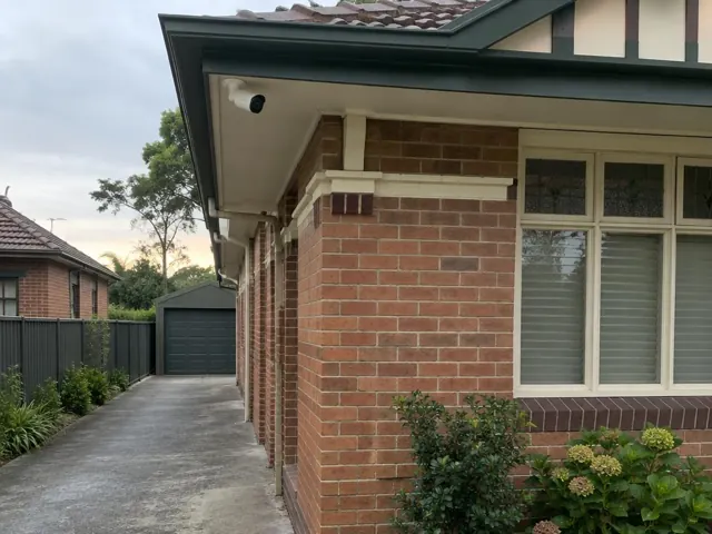 CCTV camera mounted under a brick house eave beside a driveway in Waitara